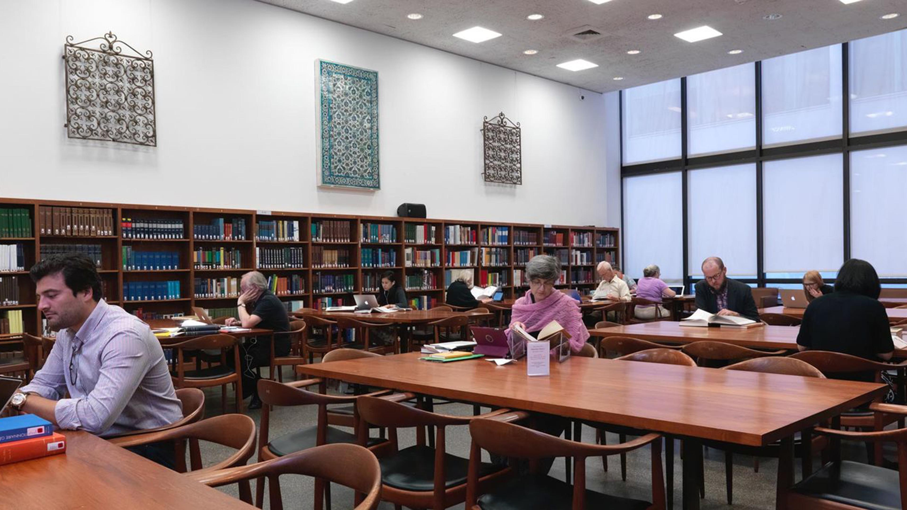 Individuals reading at wooden tables in a library with floor-to-ceiling windows on the right wall. 
