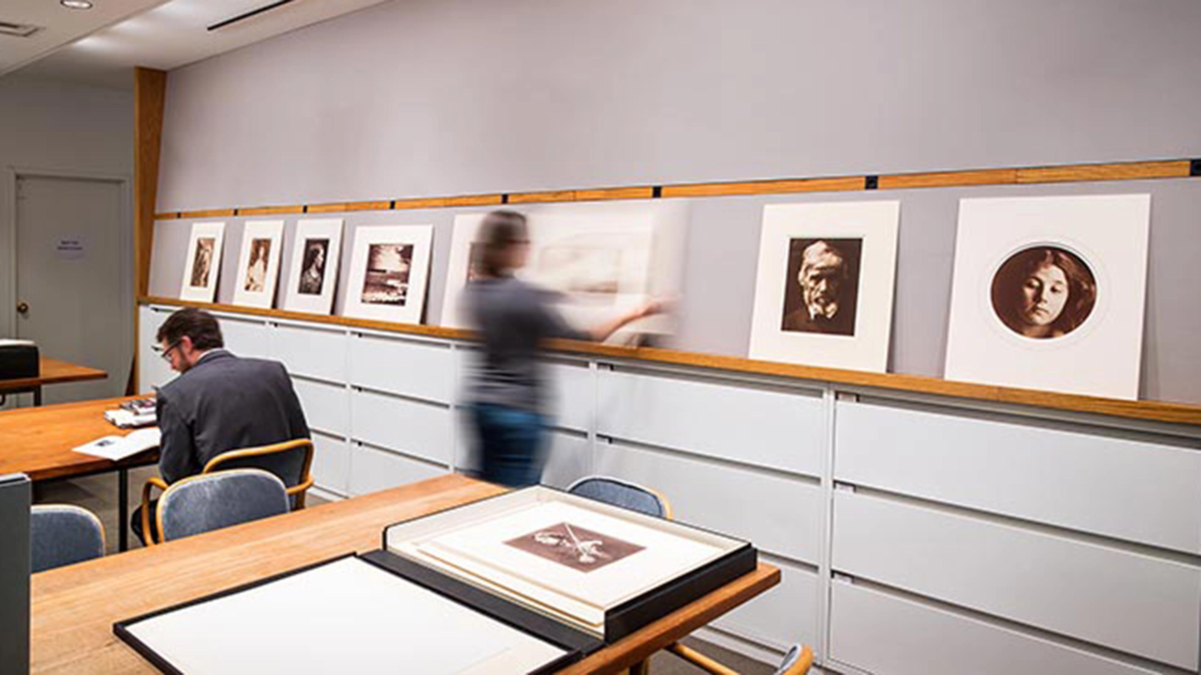 An office with blond wood tables and chairs, and against the back wall, low filing cabinets with a picture rail above for displaying photographic works of art