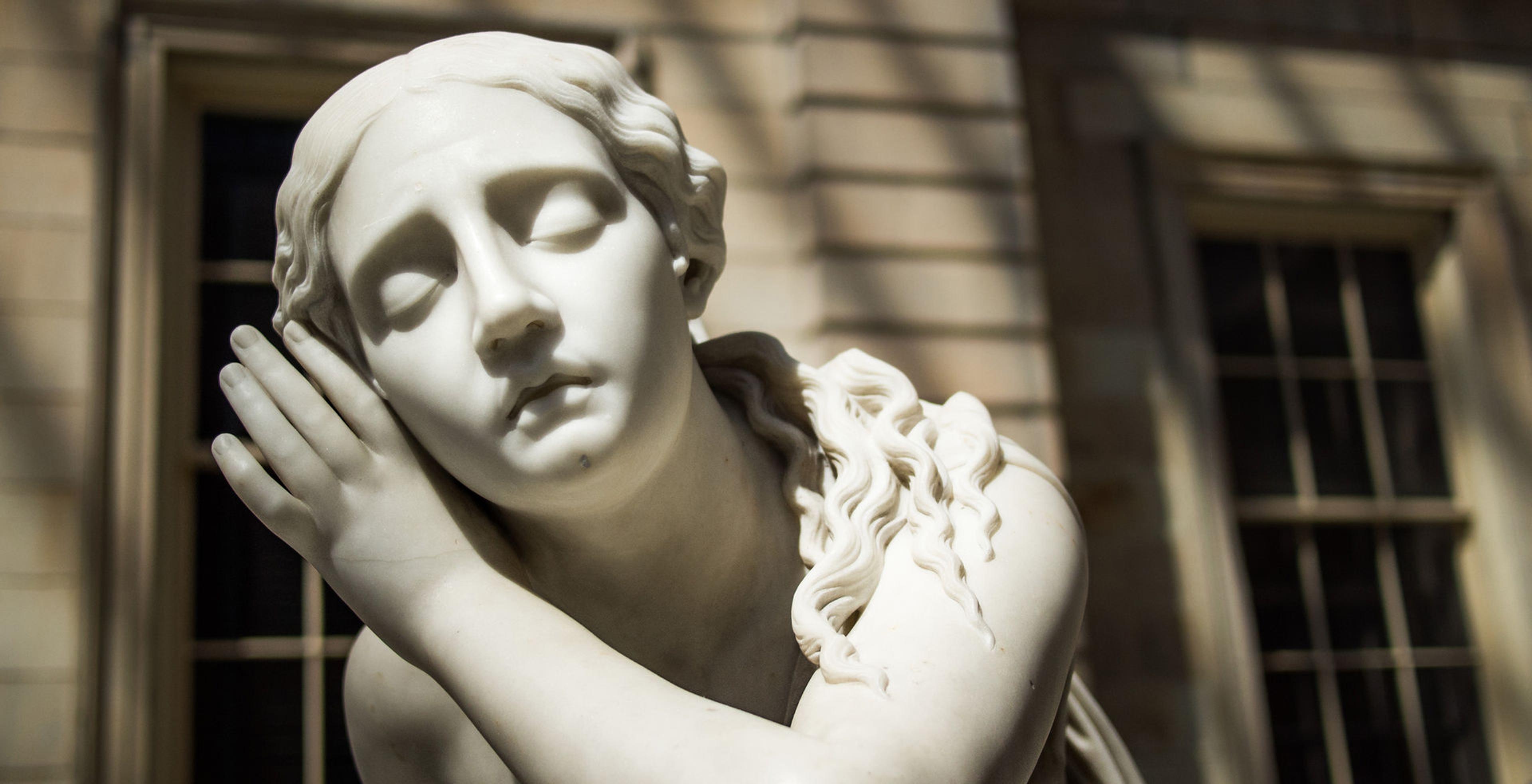 Close-up of the marble statue of Nydia, The Blind Flower Girl of Pompeii, made by Randolph Rogers, from her above her shoulders showing a young girl with closed eyes and a hand cupped around her right ear in a gesture suggesting it aids her hearing. Nydia’s face is directly facing the camera. The sculpture is in the American Wing Engelhard Sculpture Court at The Met, a skylit space with direct, dramatic natural light.