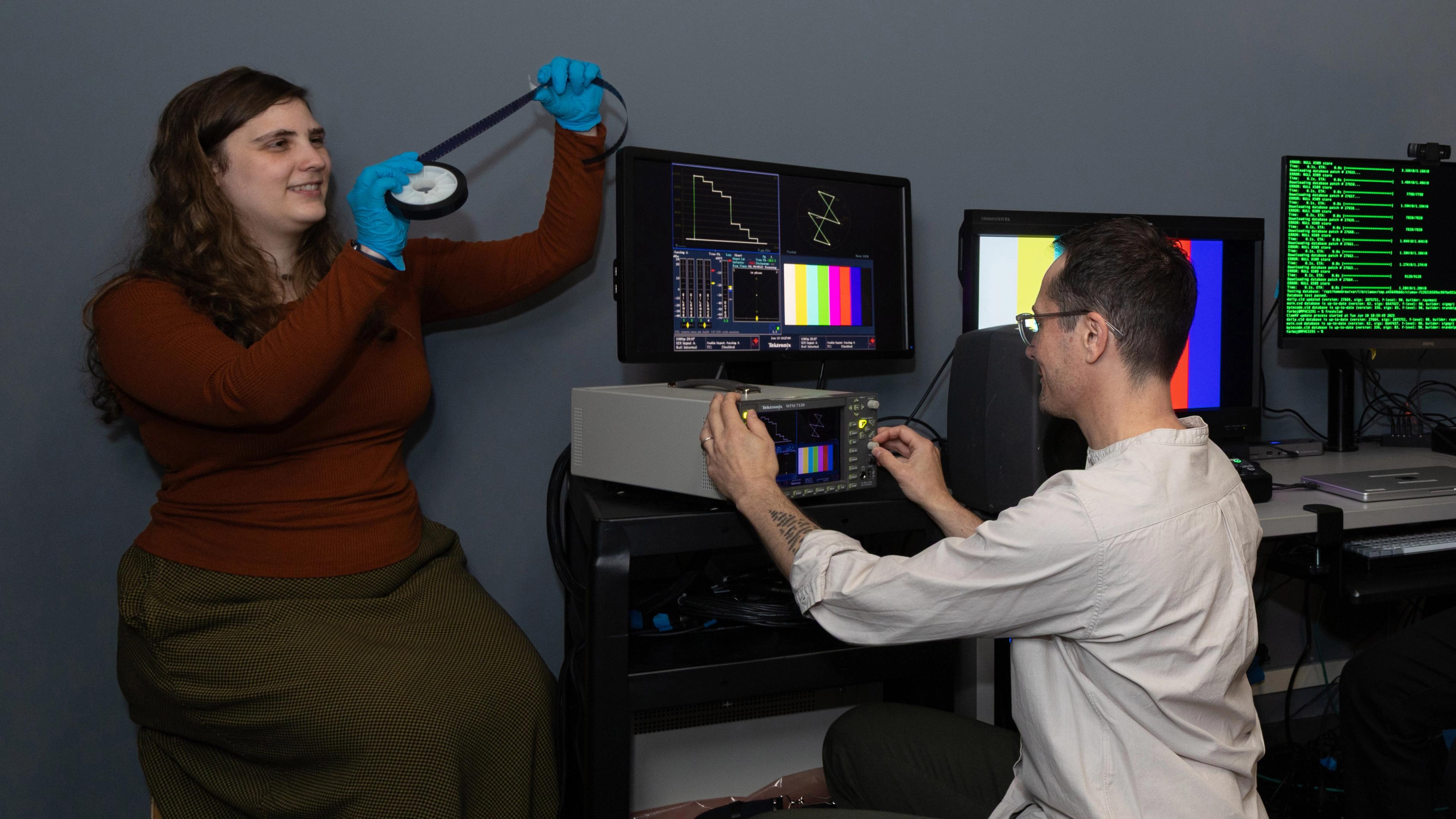 Two people work in a media lab; one handles a film reel and one operates electronic equipment.
