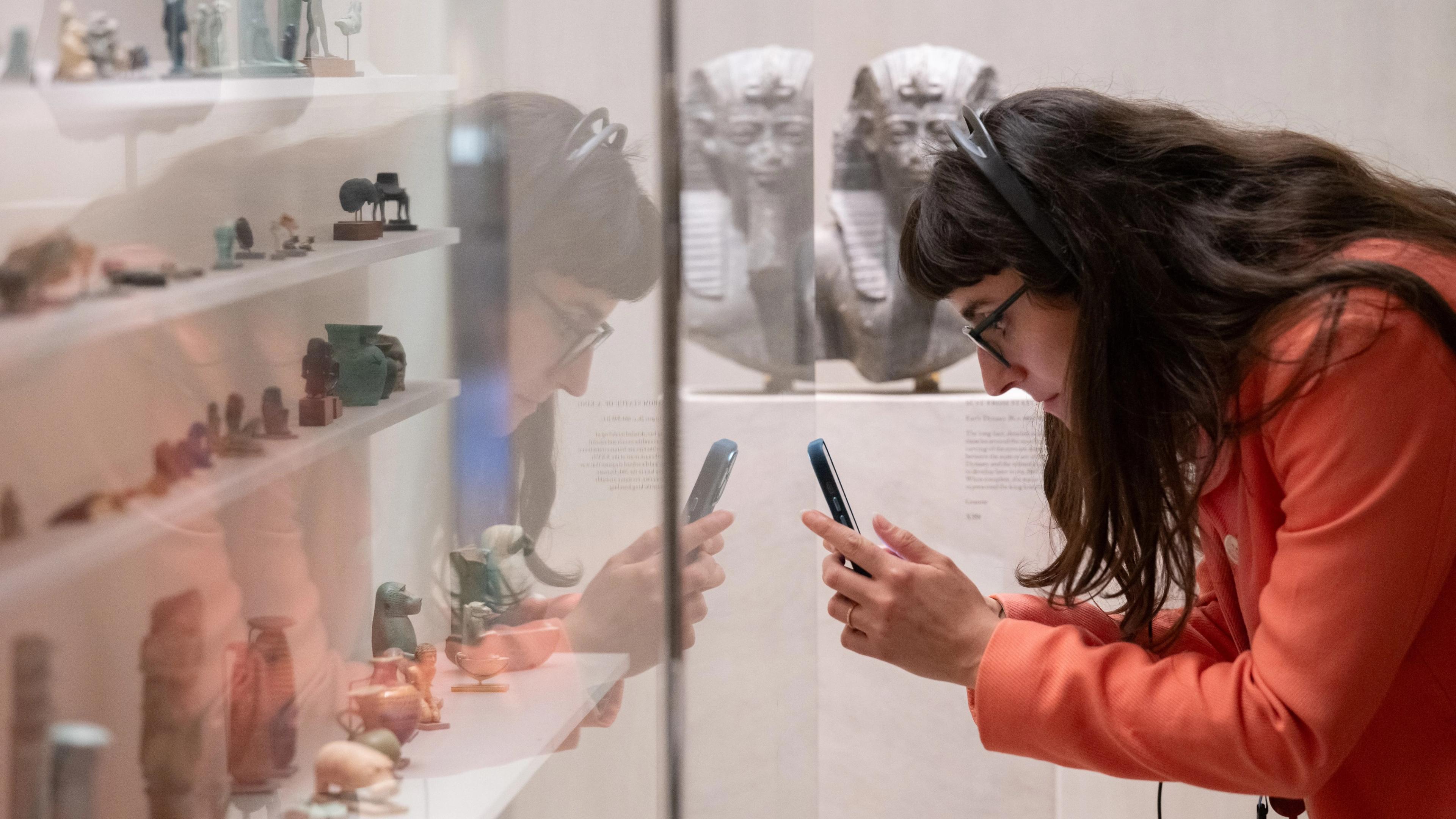A woman in glasses and a bright orange coat closely examines and photographs small Egyptian artifacts displayed in a museum case, with larger Egyptian sculptures visible in the background.
