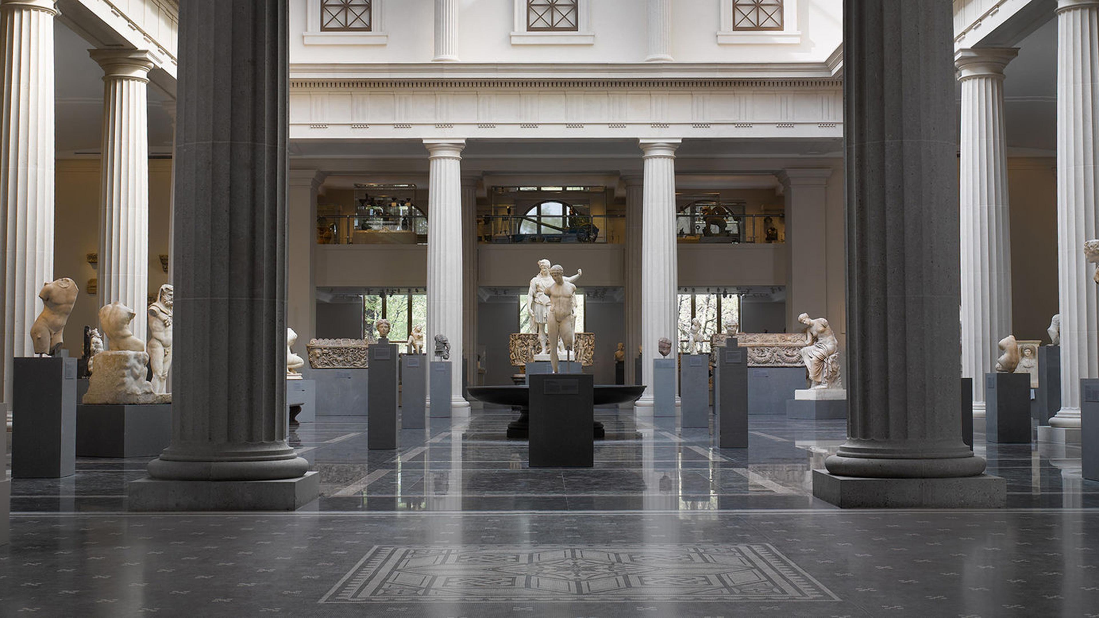 A view of the Greek and Roman Galleries in soft daylight with no visitors. The contrasts of the black floor and pedestals and white stone statues and columns creates a dramatic image.