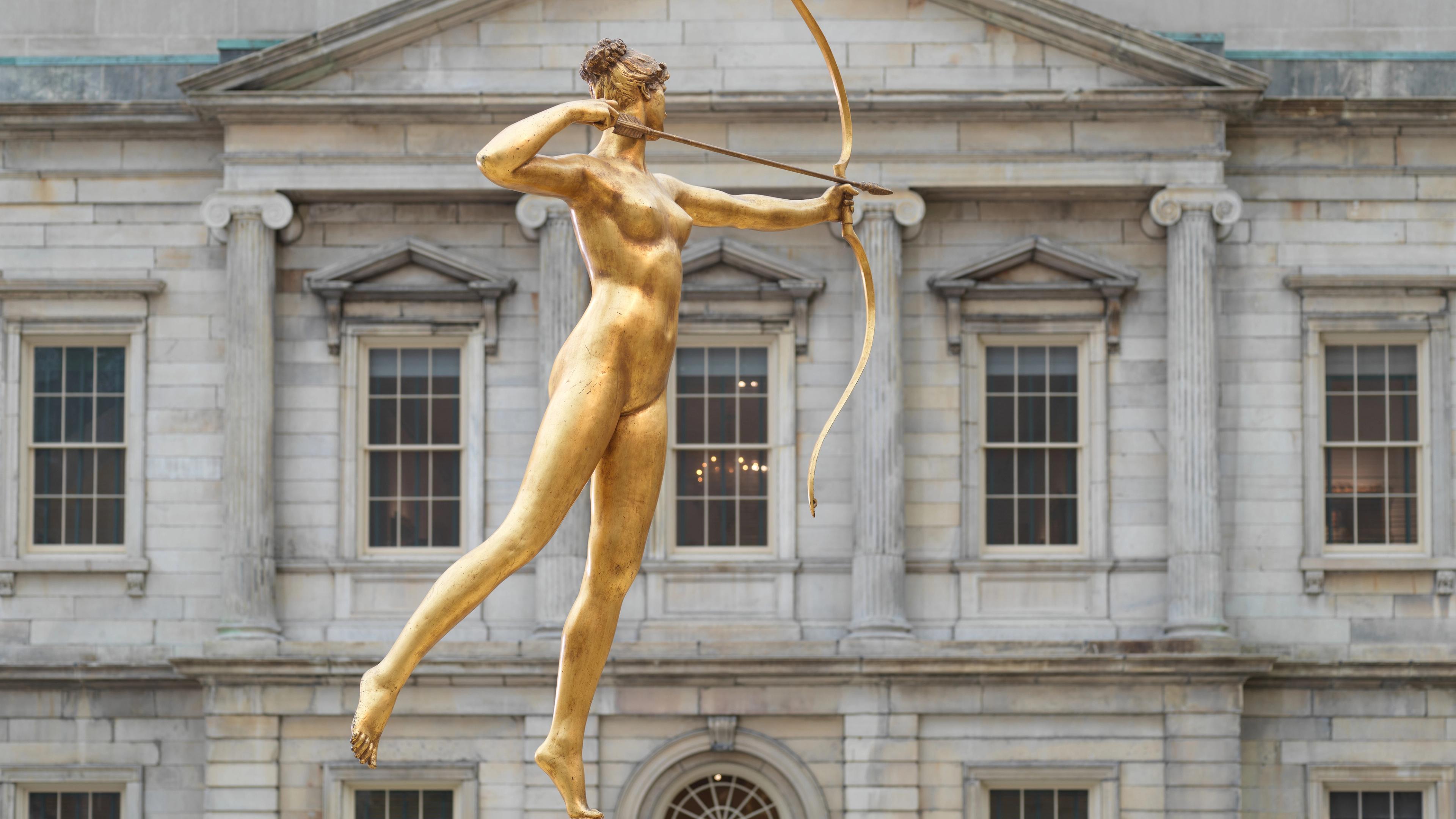 Gold statue of a nude woman, pointing a bow and arrow in the direction of a stately gray facade.