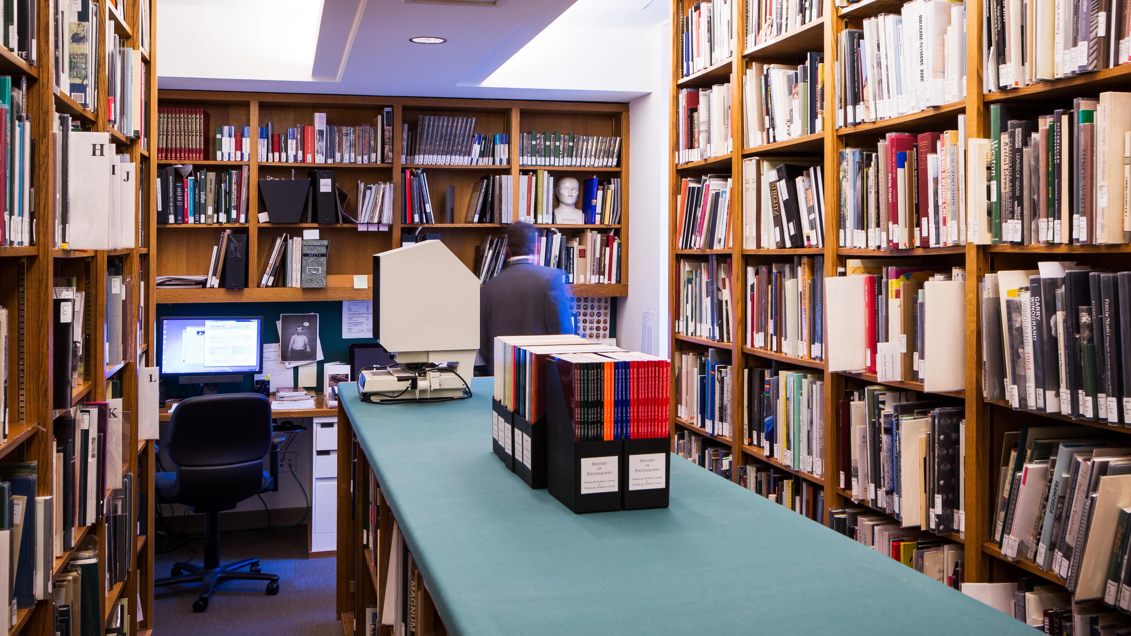 A small library with floor to ceiling wooden shelves filled with books and files and long green table down the center of the space. A person is browsing the shelves in the background next to a computer and microfiche reader.