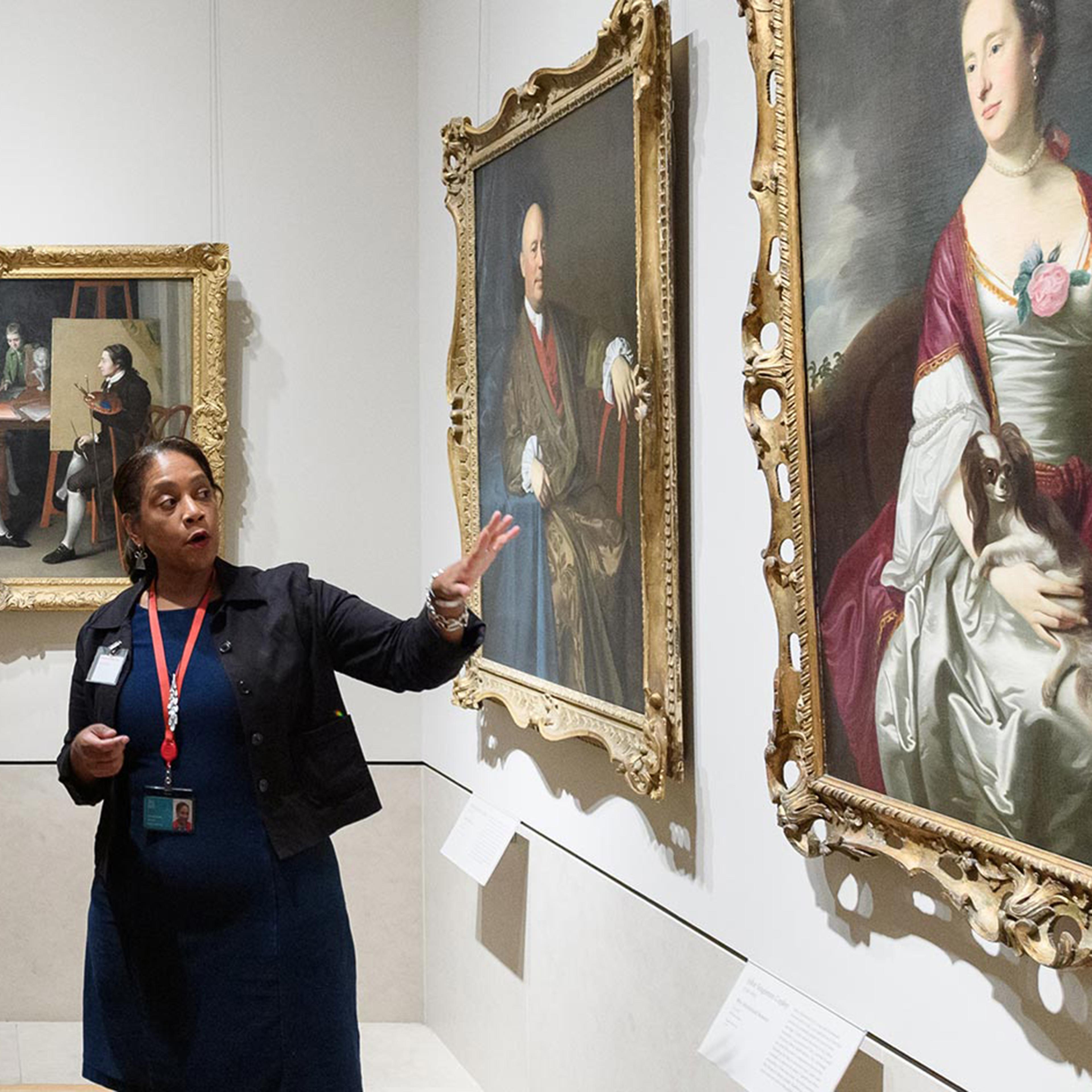 A volunteer leads a group of visitors on a tour in the American Wing. The volunteer is gesturing towards and discussing an oil painting in The Met collection.