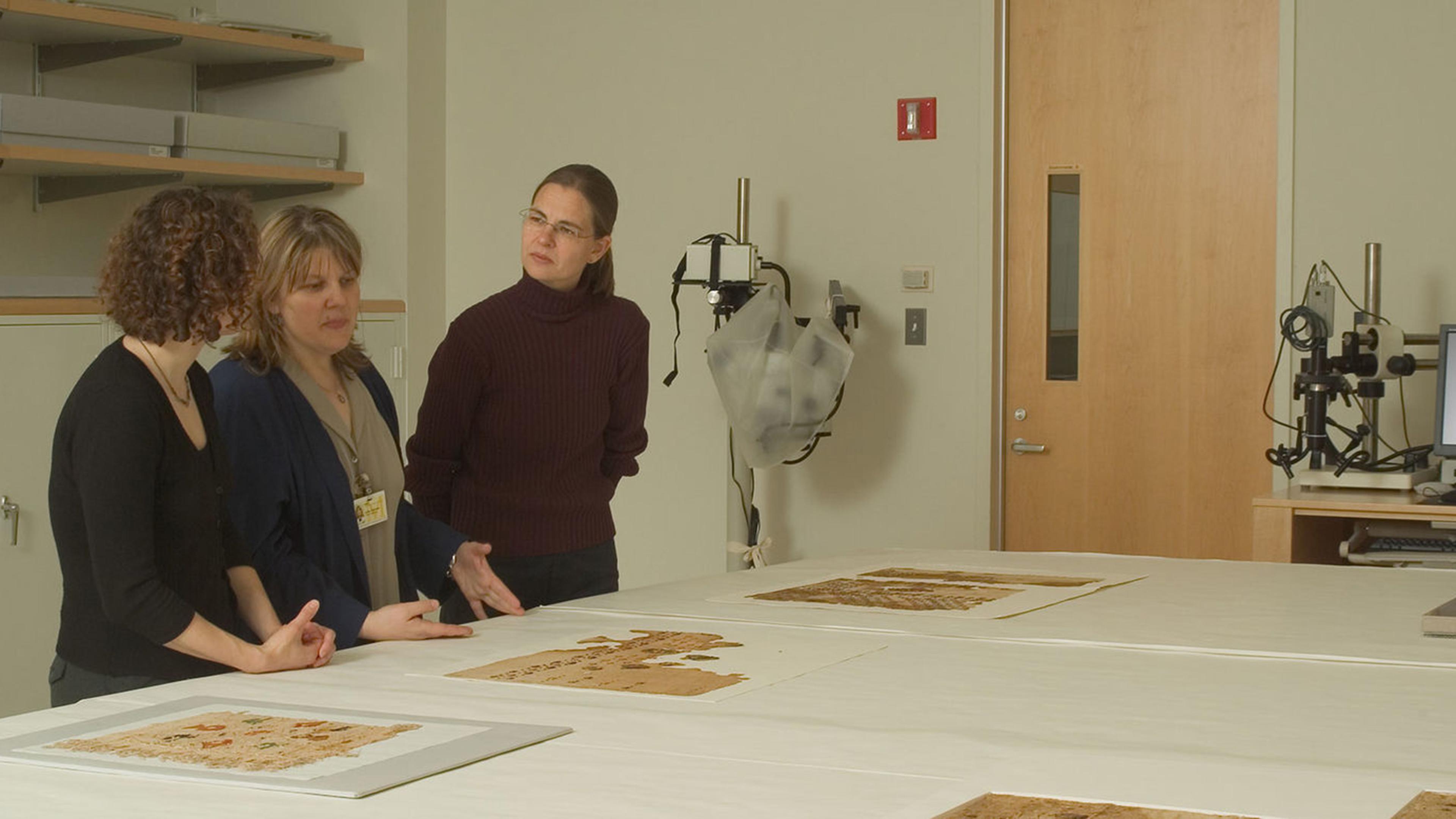 Three individuals discuss textile pieces displayed on a table in a conservation lab with a computer setup and other equipment in the background.