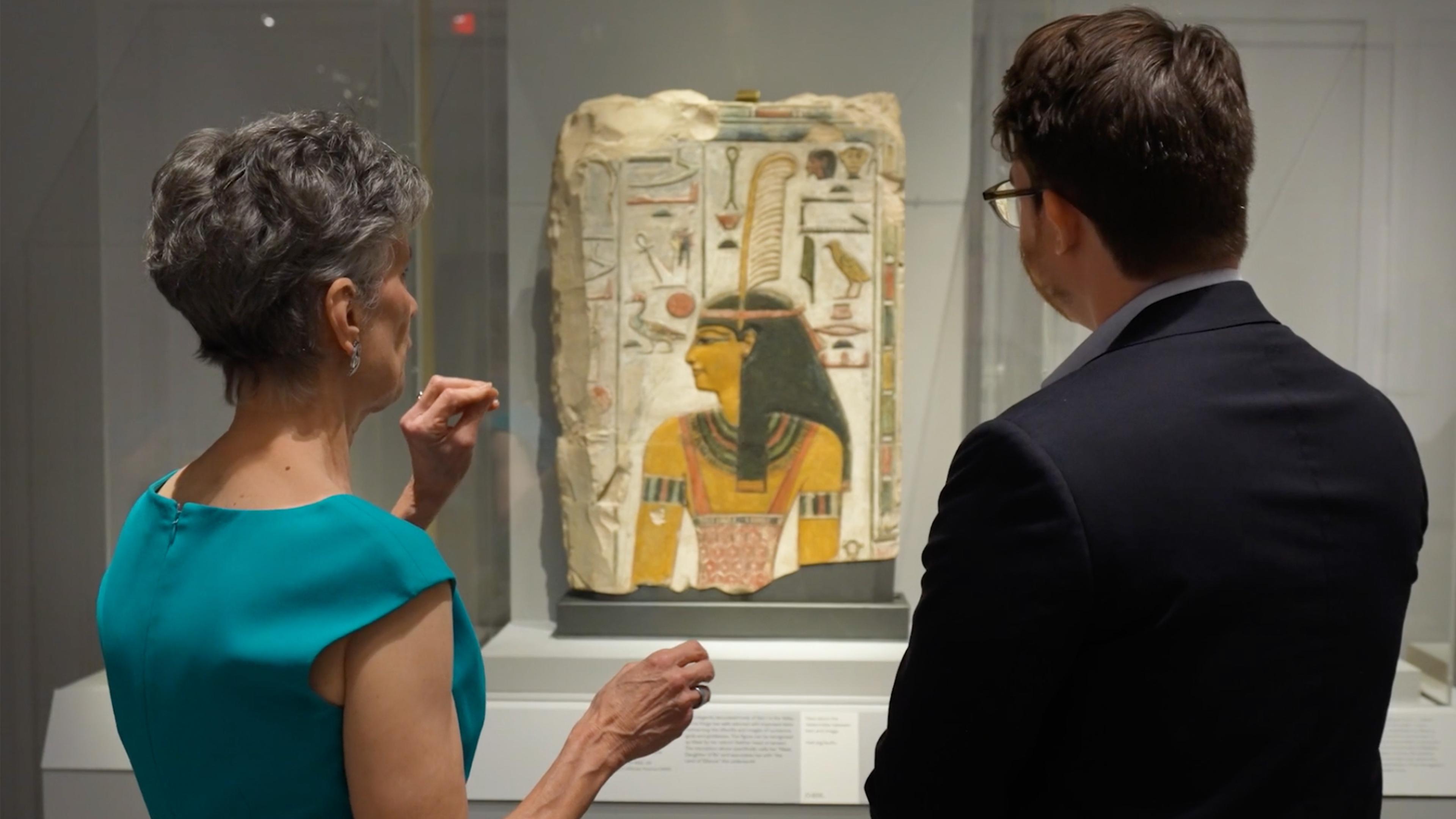 Two individuals—a woman wearing a teal-colored shirt and a man dressed in all black—stand with their backs to the camera while observing an ancient Egyptian object.