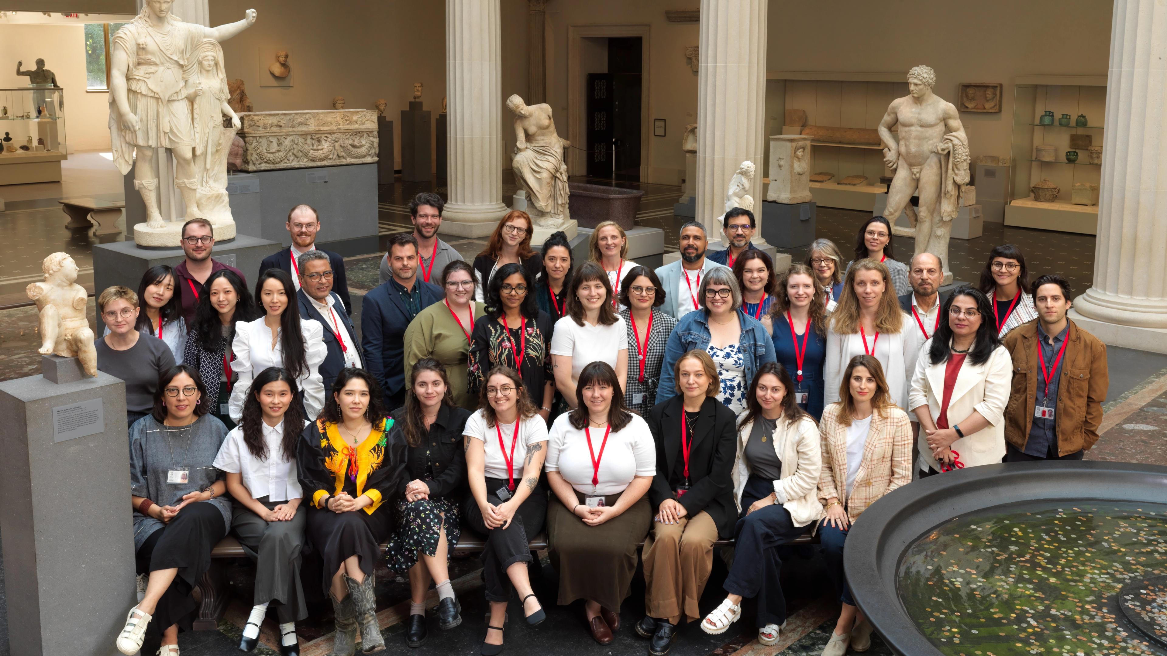 A large group of people pose for a photo inside a museum gallery with classical Greek statues and columns in the background.