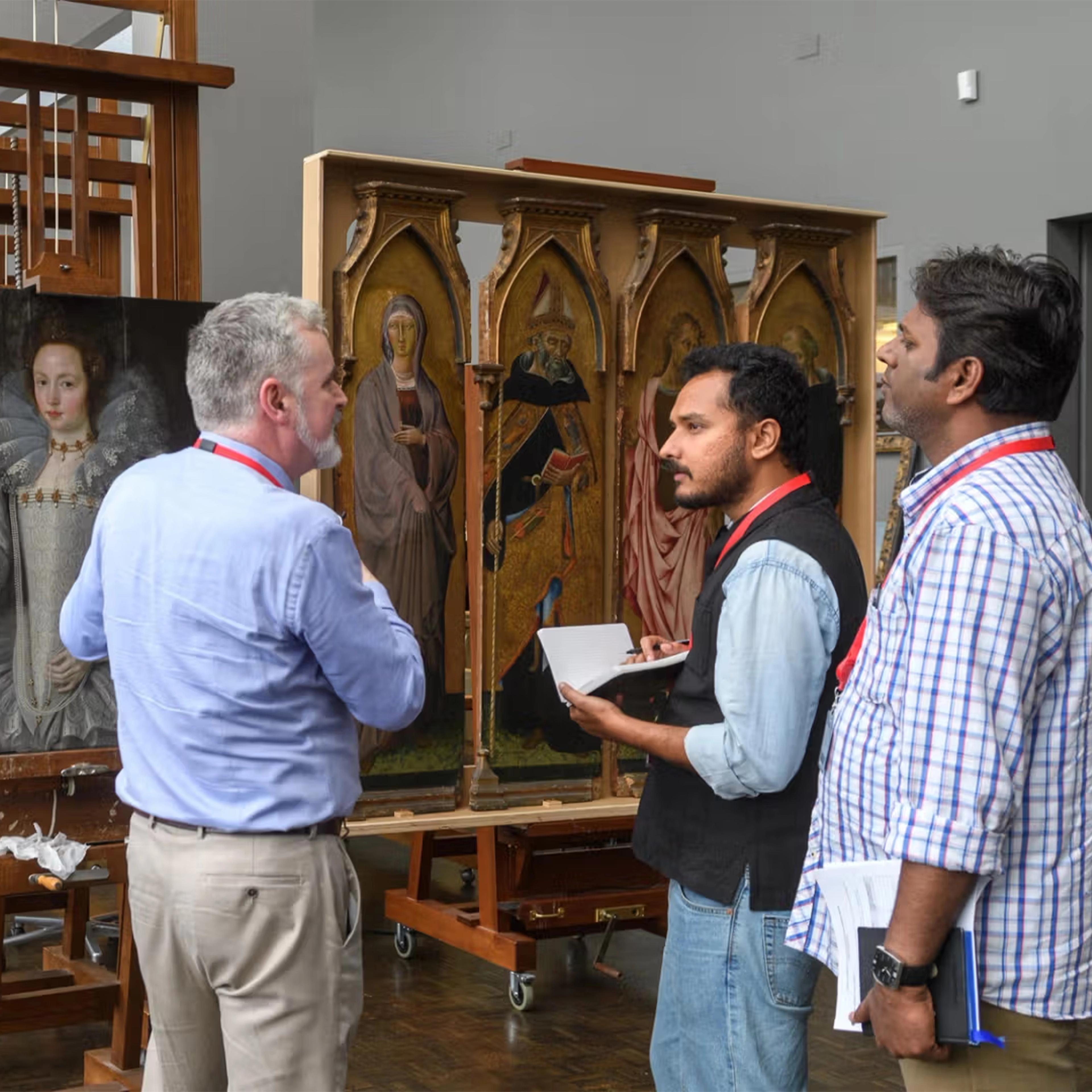Three men stand in a painting restoration workshop, examining large paintings on easels. One man in the background is in a light blue shirt and khaki pants, and is seen from behind. The other two face the artwork; one with a notebook, the other listening. They are surrounded by framed portraits paintings and altarpieces.