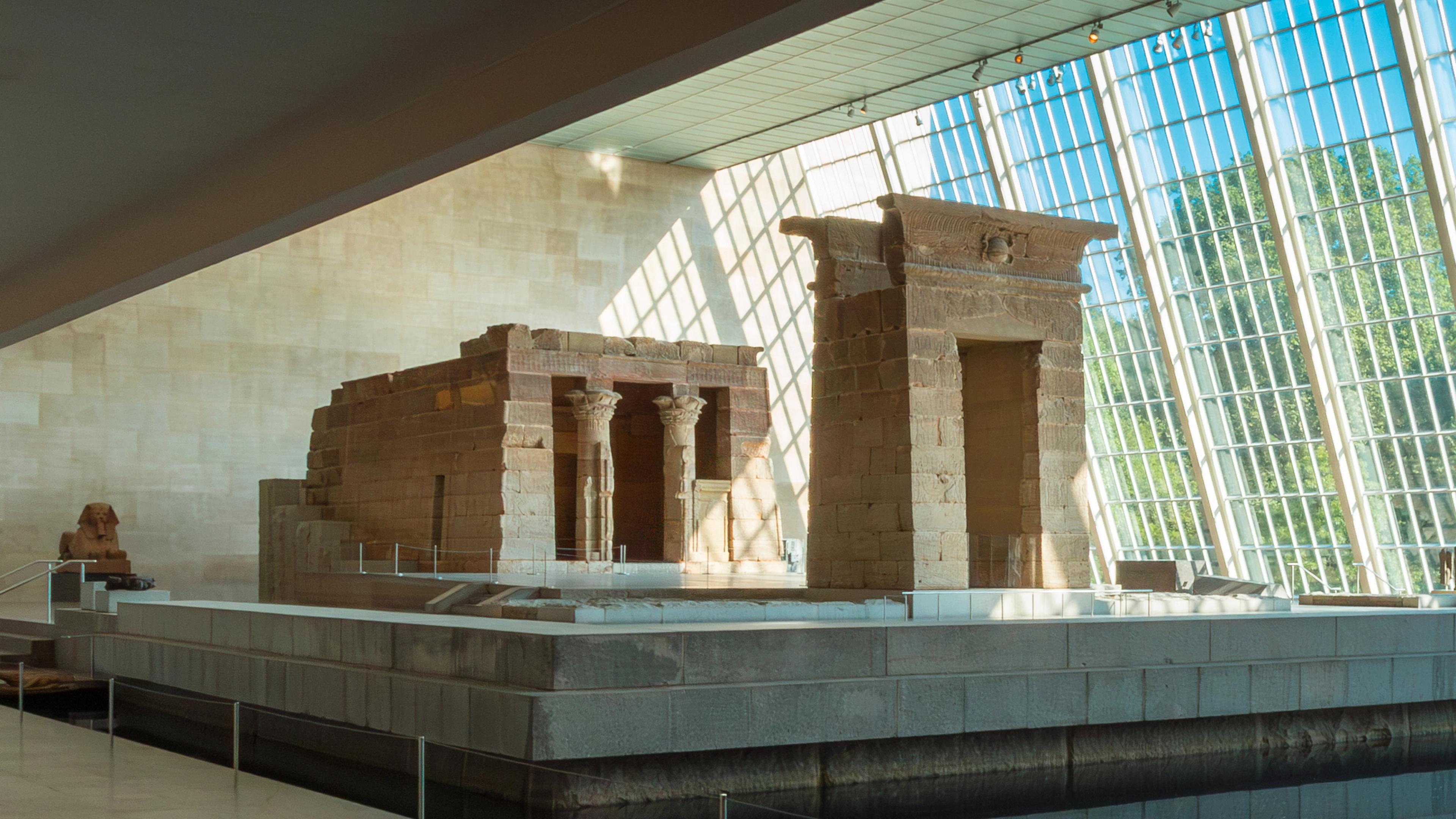 An ancient Egyptian temple structure displayed inside a museum, surrounded by a water with sunlight shining through a wall of glass windows. 