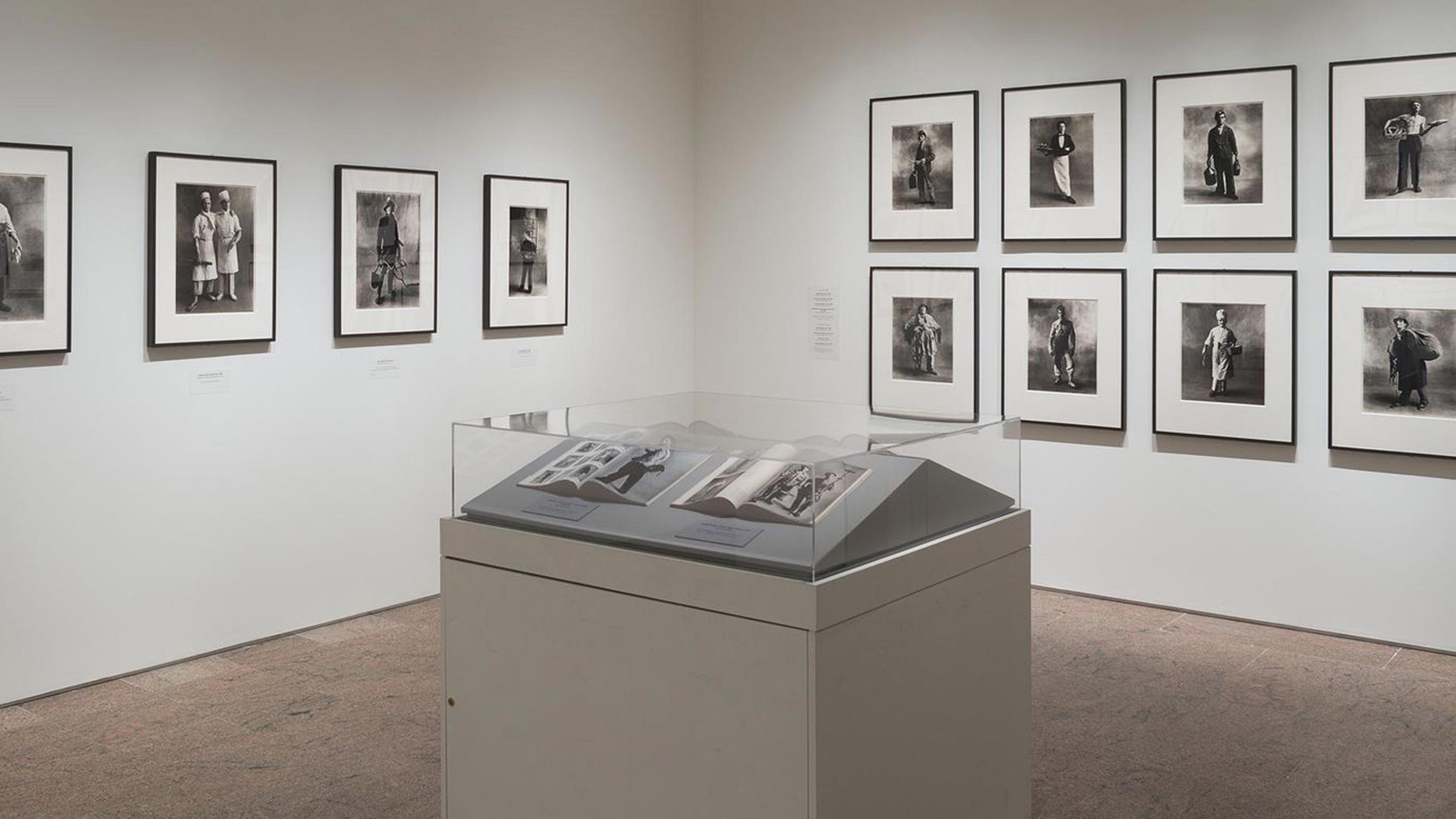 A gallery room with black-and-white portrait photographs on the walls and a display case containing open books in the center.