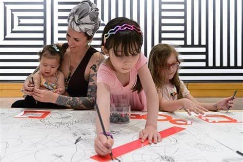 A young tattooed woman wearing a headscarf is making art with a baby on her lap. Two other toddlers are seated next to her making arts and crafts.